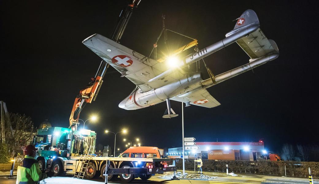 Die legendäre Vampire DH-100 wird vom Stahlgerüst gehoben, das in der Mitte des Flugplatz-Kreisels steht, und mit Gurten gesichert auf den Lastwagen gestellt.